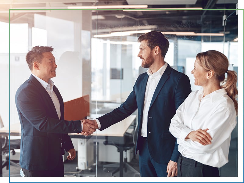 Asiatisch und europäisch gelesene Personen geben sich in einem hellen Büro die Hand als Symbol als globale Wirtschaftsbeziehungen durch SIBA GLOBAL